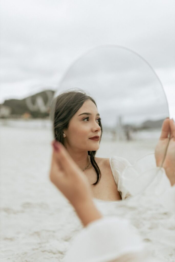 Reflective portrait of a woman holding a mirror on a beach, gazing thoughtfully.